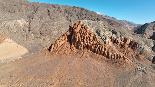 An aerial view focusing on a uniquely eroded, reddish-orange conical rock formation rising from a dry, arid plain against dark, barren mountain slopes