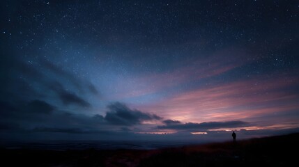 Fototapeta premium A solitary figure observes a star filled night sky painted with vibrant twilight hues and dramatic clouds