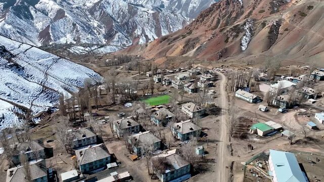 Wide aerial view of the isolated former Soviet uranium mining village nestled at the base of steep, barren mountains with colorful rock layers and snow patches