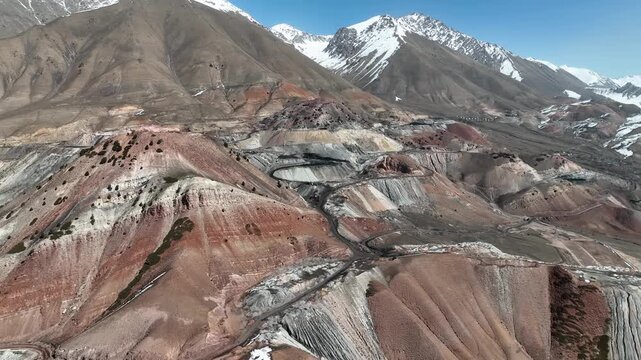 Aerial view of a massive terraced open pit or quarry in a colorful mountain landscape, featuring steep switchback roads and exposed multi-hued rock strata