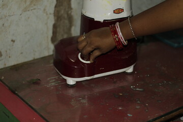 Hand of an Indian woman with bangles operating a kitchen appliance, symbolizing domestic work, routine, and the growing use of modern tools in home cooking.