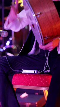 Seated musician playing darbuka with vibrant lighting, Onstage percussionist executing rhythmic patterns on darbuka amidst colorful lighting and equipment visible