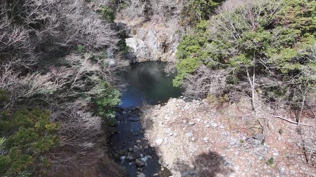 Aerial descent from the shaded forest to the Kido River waterfall basin. Stunning close-up of rocky cliffs and flowing water, ideal for adventure, nature, or outdoor video projects.