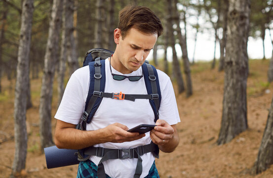 Confused hiker using smartphone for navigation, searching lost signal in pine forest. Man with backpack checking mobile phone with cellular network or battery problems, looking at connection failure