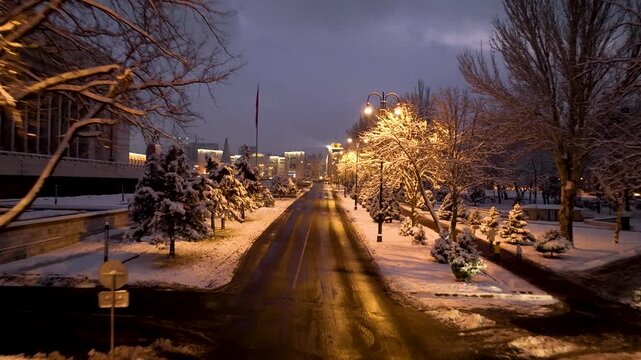 Nighttime view down a major avenue showing snow-covered trees, wet asphalt reflecting streetlights, and the illuminated Manas Monument and Historical Museum