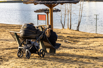 A tranquil lakeside scene with a baby stroller and two people on a wooden bench in an autumn park. Relaxed, family time, family-friendly outdoor moment.