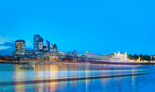 Panoramic view Tower of London and modern skyline on Thames river with motion speed light of passing ferry in the foreground at twilight blue hour - London, United Kingdom