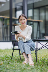 Asian woman looking at smartphone in hand writing in notebook and sitting at wooden table. Study or Working Outdoor.
