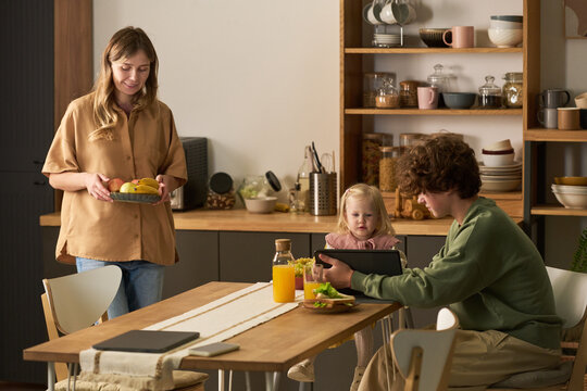 Caucasian young adult woman carrying plate of fruit, while Caucasian boy using digital tablet with Caucasian toddler girl sitting at dining table in modern kitchen