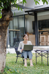 A young adult woman works on a laptop in a modern office. Sunlight streams through large windows, highlighting the greenery outside and creating a bright, serene atmosphere.
