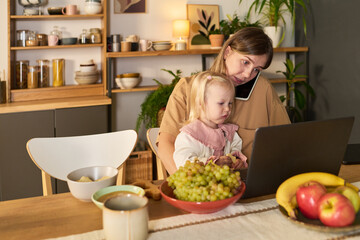 Caucasian young adult woman multitasking with smartphone between ear and shoulder, while holding Caucasian toddler girl on lap and working on laptop at kitchen table