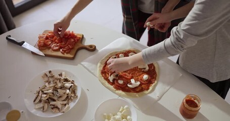 Top view of woman with her daughter putting ingredients on pizza while cooking in cozy kitchen at home. Cooking family time spending time at home
