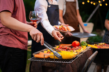 An Asian woman with friends grilling BBQ skewers, corn, and sausages outdoors. They toast with wine amid glowing string lights, creating a joyful and warm evening barbecue atmosphere