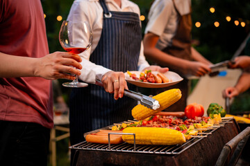 An Asian woman with friends grilling BBQ skewers, corn, and sausages outdoors. They toast with wine amid glowing string lights, creating a joyful and warm evening barbecue atmosphere