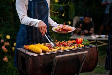 An Asian woman grills assorted barbecue skewers with meat, bell peppers, pineapple,corn outdoors. The backyard setting captures a cheerful summer cookout filled with color, warmth,delicious aromas