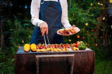 An Asian woman grills assorted barbecue skewers with meat, bell peppers, pineapple,corn outdoors. The backyard setting captures a cheerful summer cookout filled with color, warmth,delicious aromas