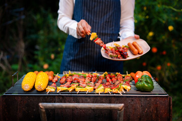 An Asian woman grills assorted barbecue skewers with meat, bell peppers, pineapple,corn outdoors. The backyard setting captures a cheerful summer cookout filled with color, warmth,delicious aromas