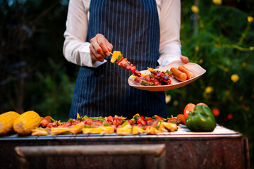 An Asian woman grills assorted barbecue skewers with meat, bell peppers, pineapple,corn outdoors. The backyard setting captures a cheerful summer cookout filled with color, warmth,delicious aromas