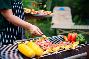 An Asian woman grills assorted barbecue skewers with meat, bell peppers, pineapple,corn outdoors. The backyard setting captures a cheerful summer cookout filled with color, warmth,delicious aromas