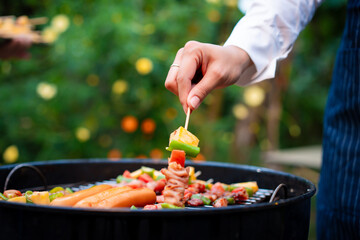 An Asian woman grills assorted barbecue skewers with meat, bell peppers, pineapple,corn outdoors. The backyard setting captures a cheerful summer cookout filled with color, warmth,delicious aromas