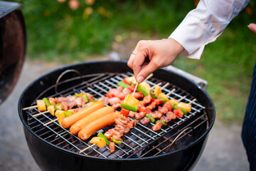 An Asian woman grills assorted barbecue skewers with meat, bell peppers, pineapple,corn outdoors. The backyard setting captures a cheerful summer cookout filled with color, warmth,delicious aromas