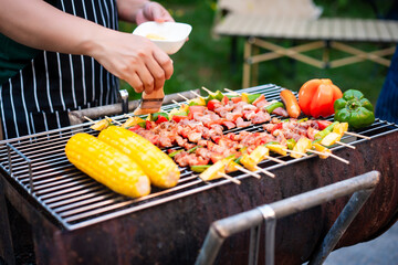 An Asian woman grills assorted barbecue skewers with meat, bell peppers, pineapple,corn outdoors....