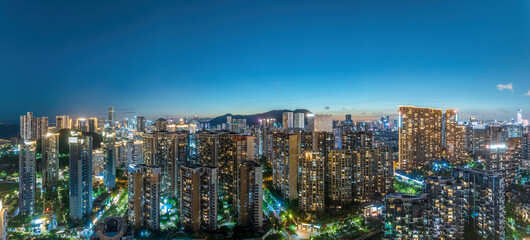 Obraz premium Wide panoramic aerial view of an illuminated modern cityscape with residential buildings and mountains in the background during twilight.