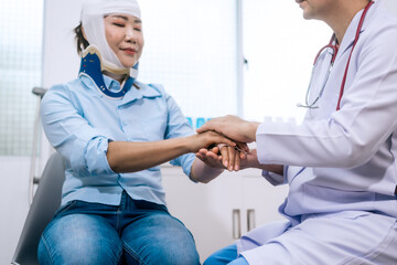 A male doctor gently examines and reassures an Asian female patient wearing a head bandage and neck...
