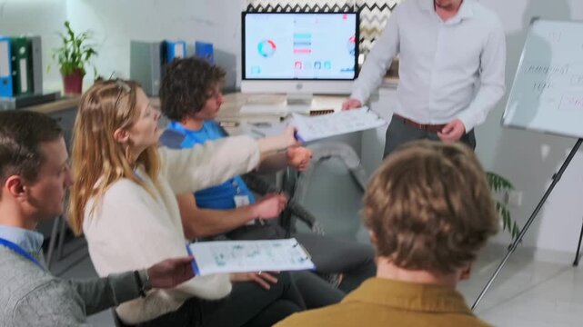 Business coach in a white shirt gathers tablets from employees seated in a circle during a corporate seminar, stacking them to return to the assistant after the group training session.