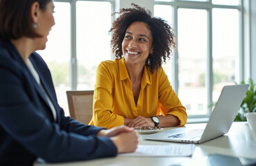 Two female colleagues have discussion in office meeting. Smiling african american businesswoman talks to her coworker at desk with laptop. Happy women working together on corporate project planning.