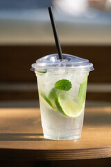 Plastic cup with lime and mint drink on wooden table in sunlight