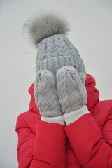 Woman in red down jacket, grey knitted hat and mittens on white background. She covered her face to protect herself from cold. Women's warm winter clothes.