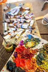 Colorful oil paint palette with messy brushstrokes, vibrant pigments, scattered tubes and finished canvas in the background. Artistic chaos as symbol of creativity and inspiration