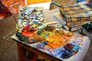 Artist creative workspace with oil paint palette, colorful brushstrokes, paint tubes, glass jar, chair and canvas in the background. Real studio atmosphere full of artistic energy.