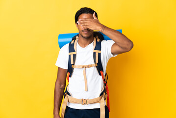 Young mountaineer man with braids with a big backpack isolated on yellow background covering eyes...