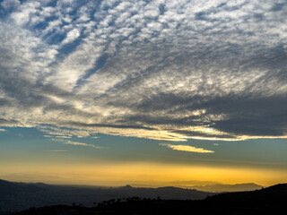 Golden sunset above Athens, Greece, featuring dramatic layered clouds spreading across the sky, casting warm light over distant hills and cityscape within the tranquil evening atmosphere