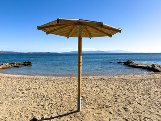 A single wooden beach umbrella stands upon the quiet sandy shore under bright midday sunlight, facing calm blue sea waters and distant mountains within a clear serene atmosphere