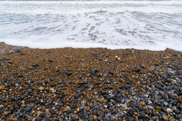 Pebble beach shoreline with white ocean waves washing over colourful stones and rocks in Cromer, UK, natural coastal seascape with foaming surf on the British seaside pebbled shore
