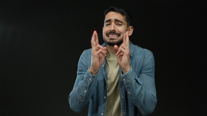 Young man with fingers crossed expressing hope in denim jacket against black background portrays anticipation and hopefulness.