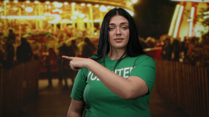 Woman volunteer making gesture at outdoor fair with bright lights and crowd, wearing green shirt, capturing curvy female empowerment and community spirit in festive setting.