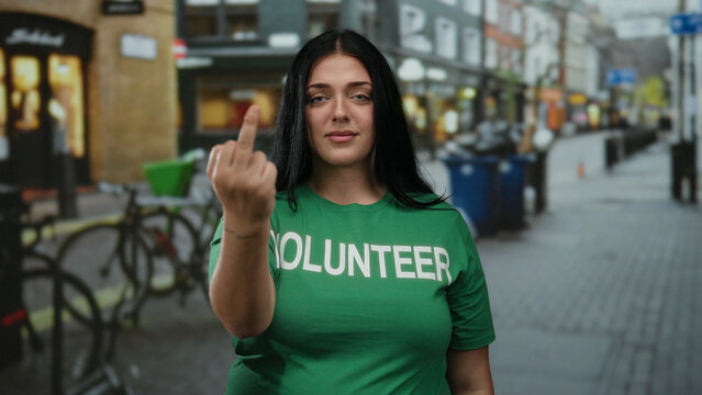 Woman in green volunteer shirt standing on city street makes unfriendly gesture, showing middle finger, with blurred urban background. - Powered by Adobe