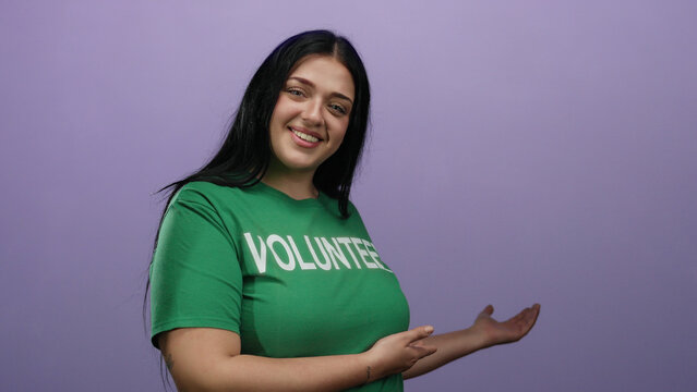 Young volunteer woman in green shirt presenting with hands against a purple wall background, showcasing a welcoming and friendly atmosphere.