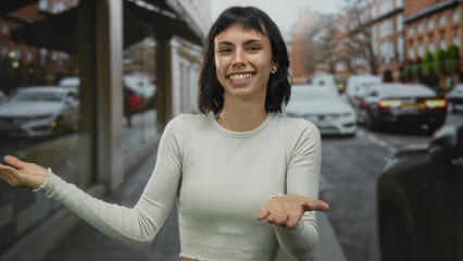 Fototapeta premium Woman in white long sleeve top shows open palms and smiles amid parked cars on urban street; welcoming.