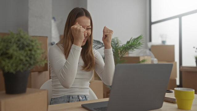 Woman typing on laptop at a wooden desk surrounded by cardboard moving boxes and potted plant in a building new home; fresh start joy.