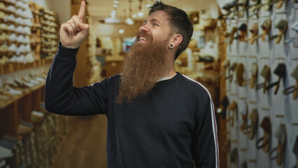 Young bearded man pointing both index fingers upward at a shoe display in an indoor shoe store with...