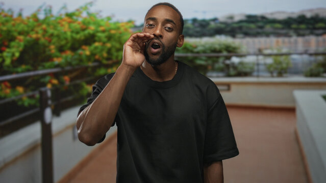 Young african american man cupping hands to mouth shouting on building balcony outdoors near railing and planters overlooking city skyline; urgent message.