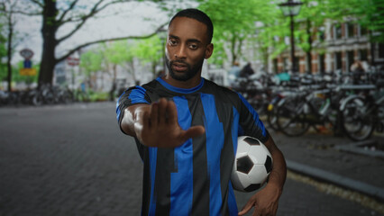 Young african american man in blue jersey holds soccer ball and shows palm out stop gesture on city street; team support determination.