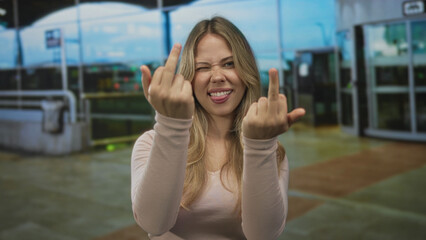 Fototapeta premium Blonde woman flips middle finger gesture with both hands at an airport terminal; defiance rebellion frustration boldness.