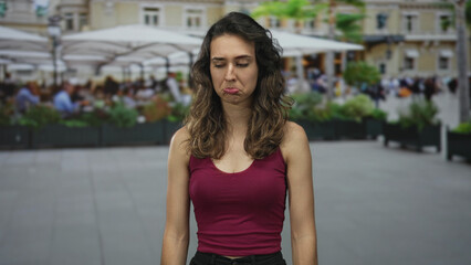 Woman in maroon tank top alone frowns with closed eyes on street caf&eacute; terrace under midday sunlight; sadness.