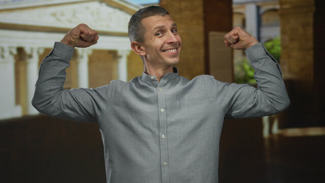 Man showing confidence by flexing arms in front of an old university building outdoor, wearing a gray shirt and expressing strength and determination in an educational setting.